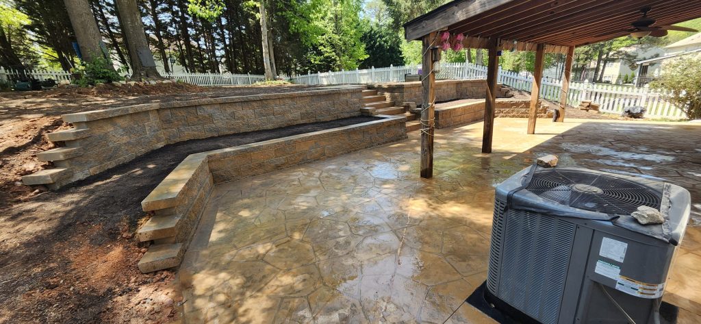 A backyard patio with stamped concrete, stone retaining walls, and a covered area next to an outdoor AC unit.