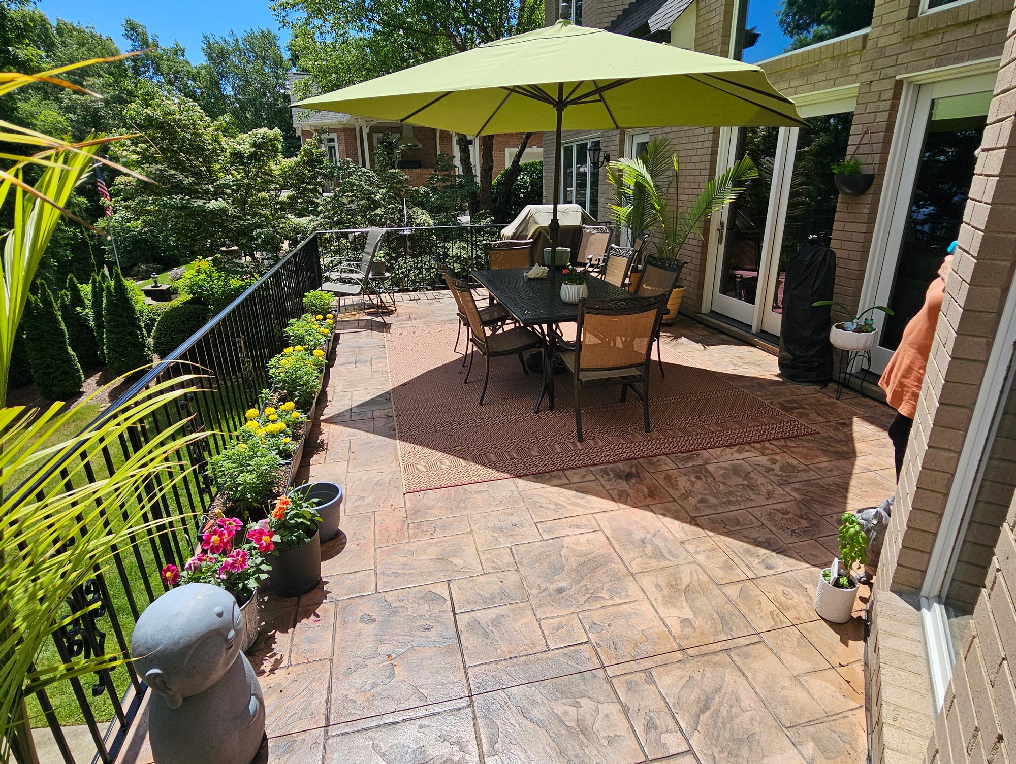 Sunny patio with table, chairs, green umbrella, potted plants, and flowers along the railing.