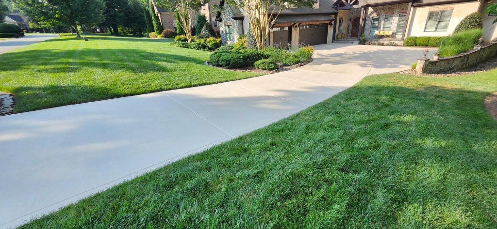 A clean concrete driveway curves toward a house with a well-kept lawn and landscaped garden.