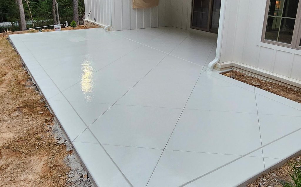 Smooth, light-gray concrete patio with subtle geometric lines beside a white house and wooded area, crafted by expert concrete services for a polished outdoor space.