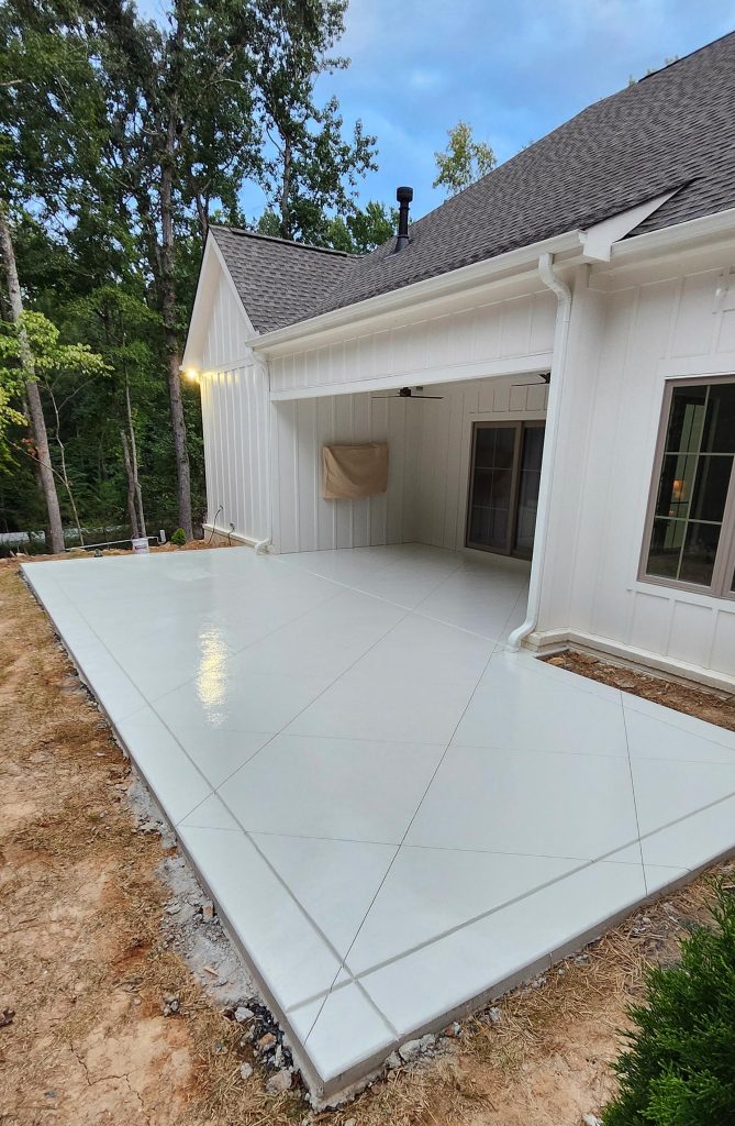 A freshly poured and sealed concrete patio behind a white house with trees in the background.