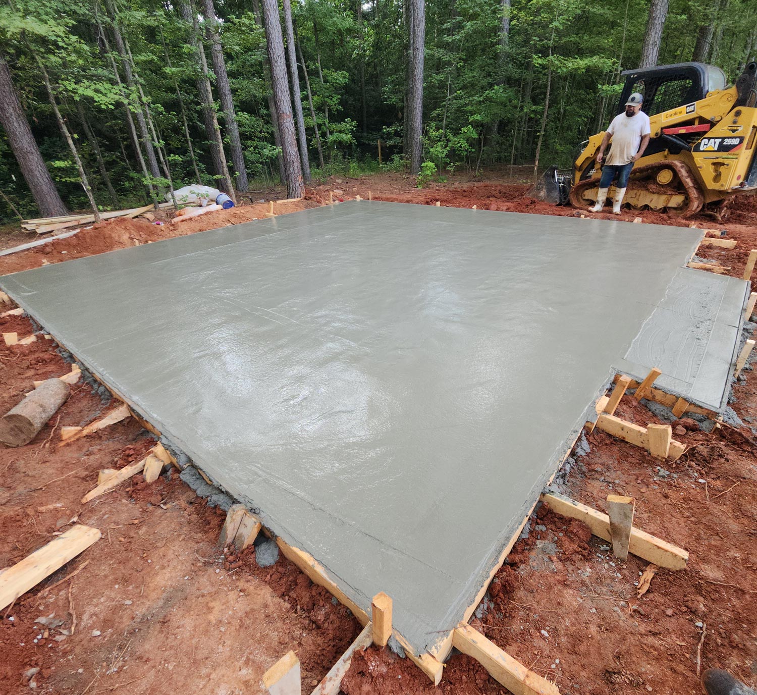 Freshly poured concrete slab curing in a wooded area, with a worker and bulldozer nearby.