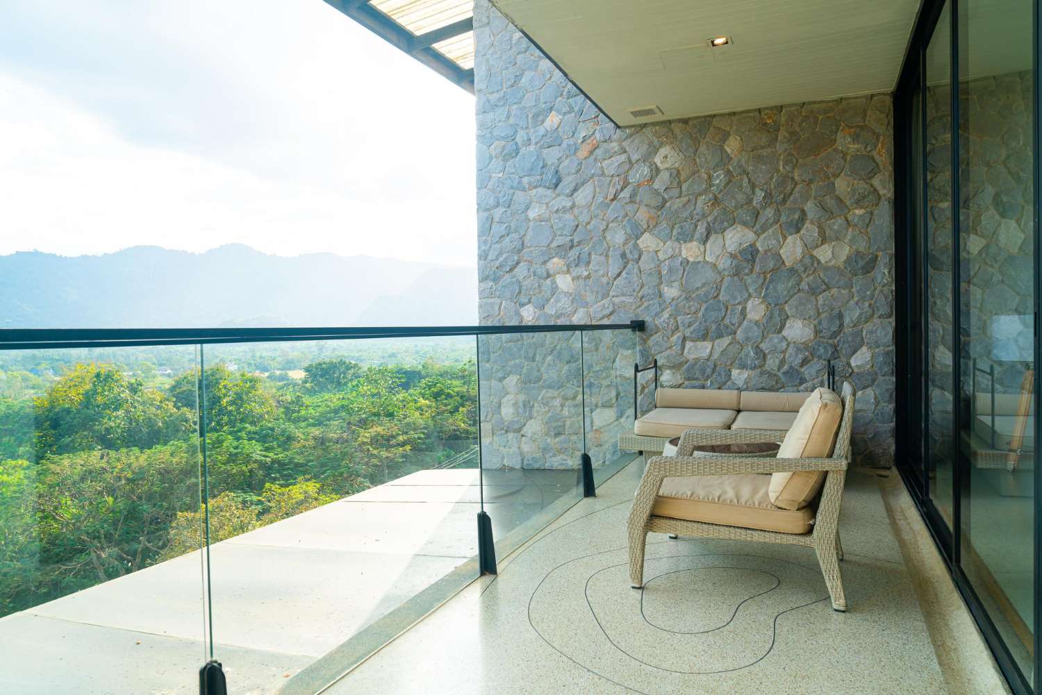 Modern balcony with stone wall, glass railing, and outdoor seating overlooking lush green trees and distant hills.