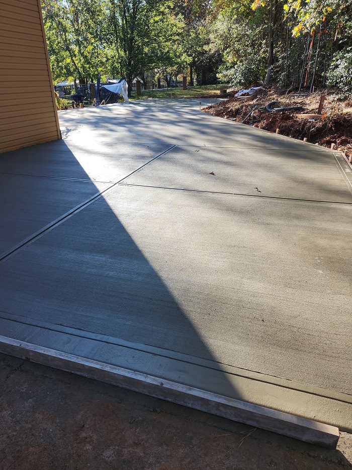 Freshly poured concrete driveway next to a house, drying in the sunlight, with trees in the background.