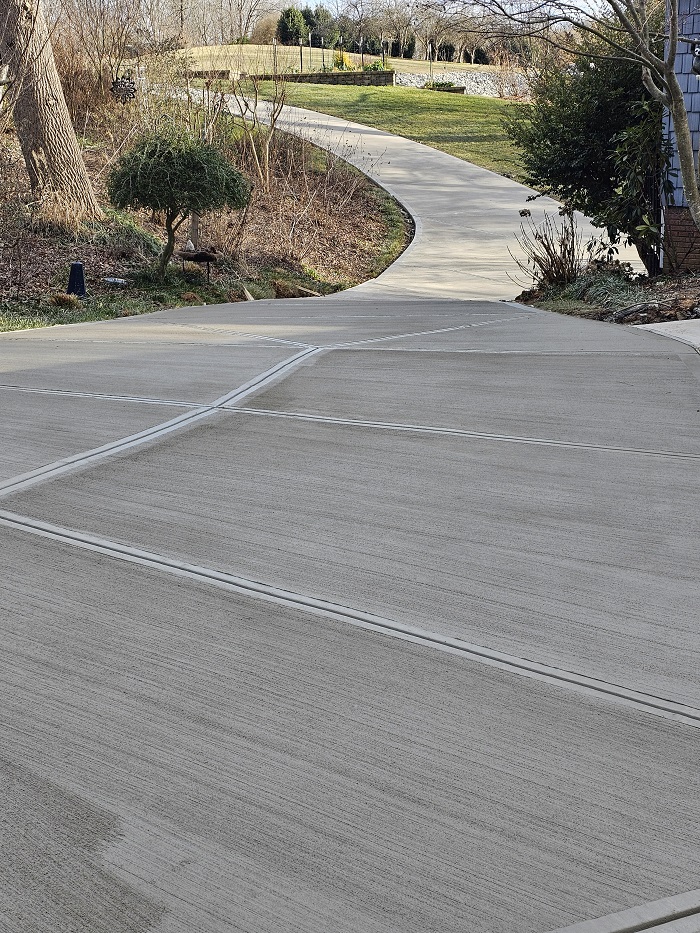A smooth concrete driveway curves into a landscaped yard with trees and bushes on a sunny day.