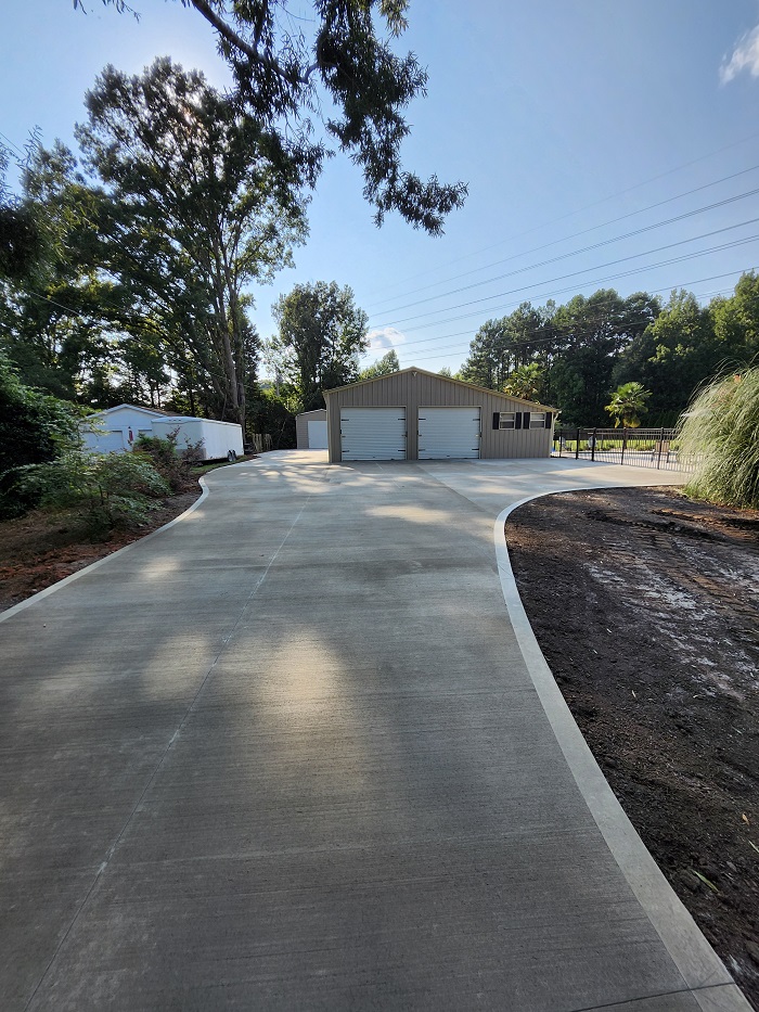 A wide, newly paved concrete driveway curves toward a large garage with three doors, surrounded by trees.