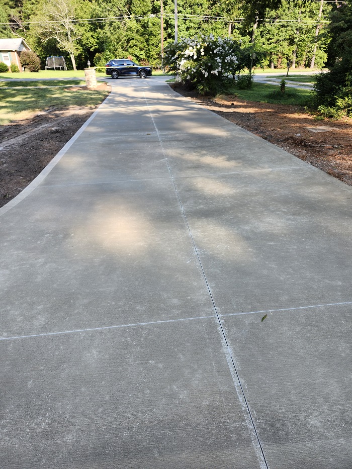 Freshly paved concrete driveway leading to a black car parked near trees and a flowering bush.
