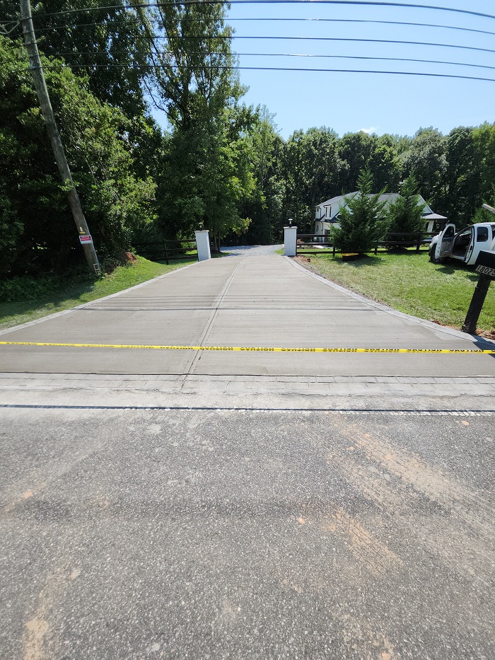 Freshly paved driveway blocked by caution tape, leading to a house with trees in the background.