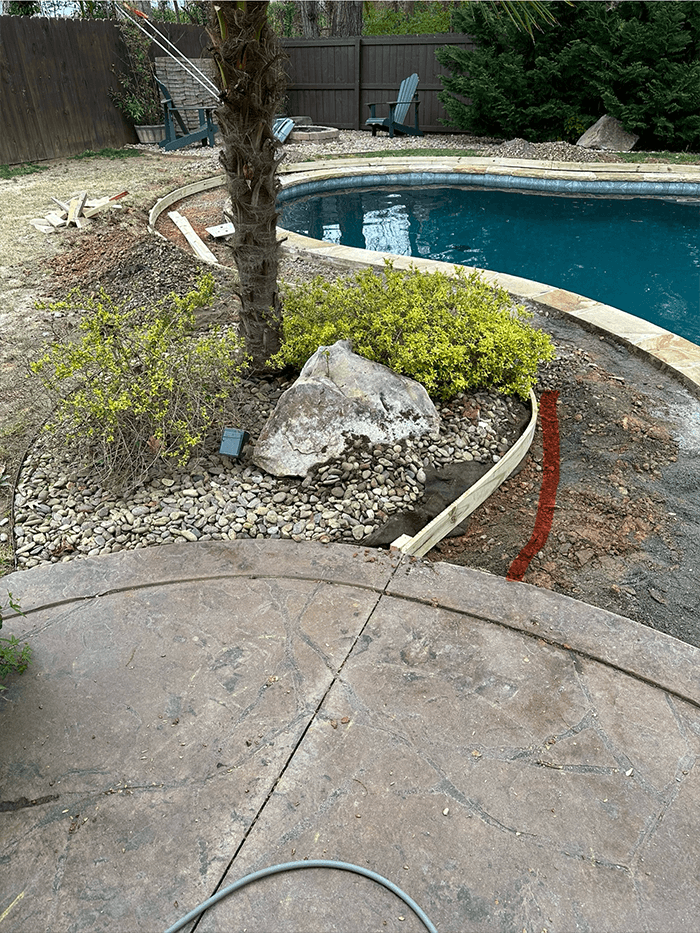 Backyard pool with concrete patio, landscaping rocks, plants, a palm tree, and a wooden garden border under construction.