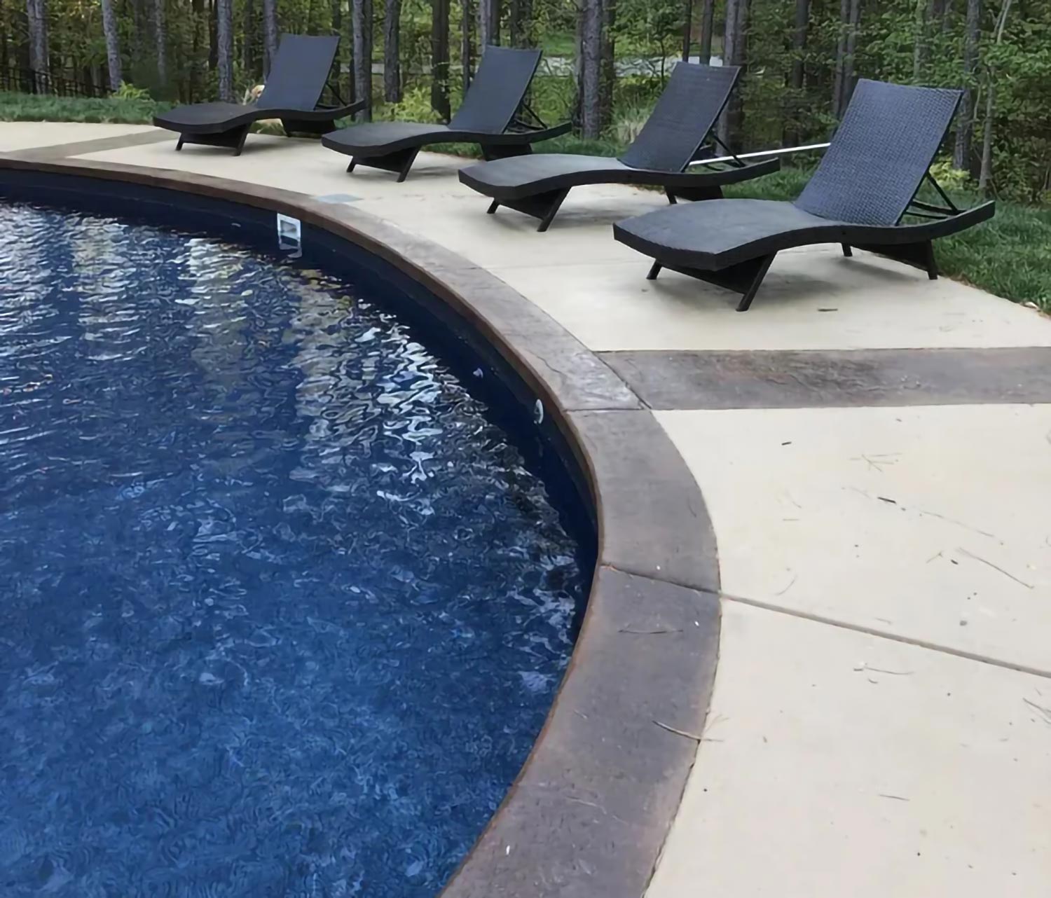 Four black lounge chairs sit beside a curved pool, surrounded by trees and a concrete deck.