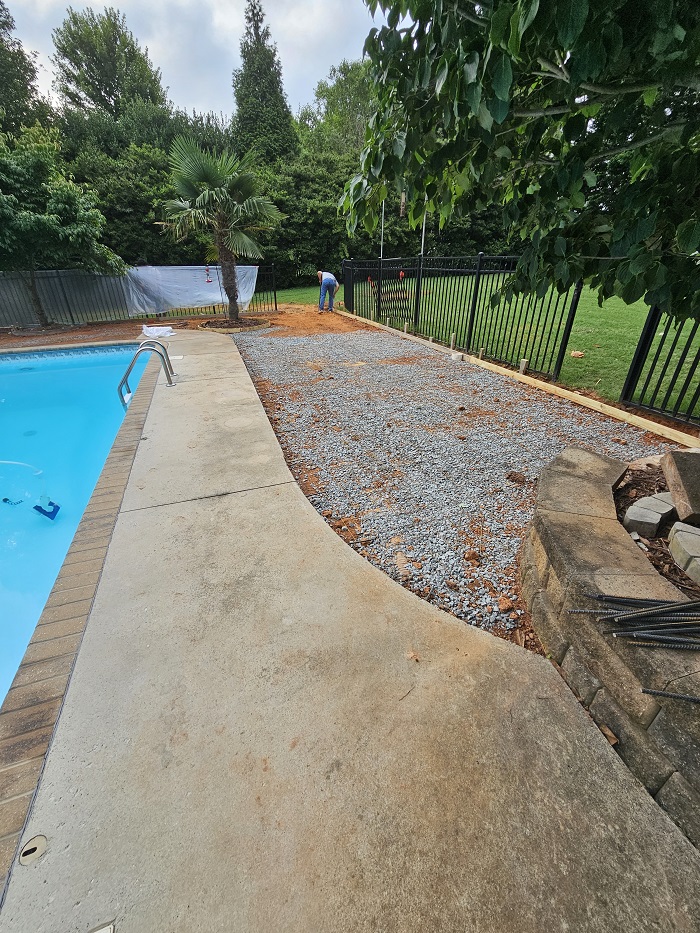 A person works near a gravel area by a swimming pool, with trees and a black fence in the background.