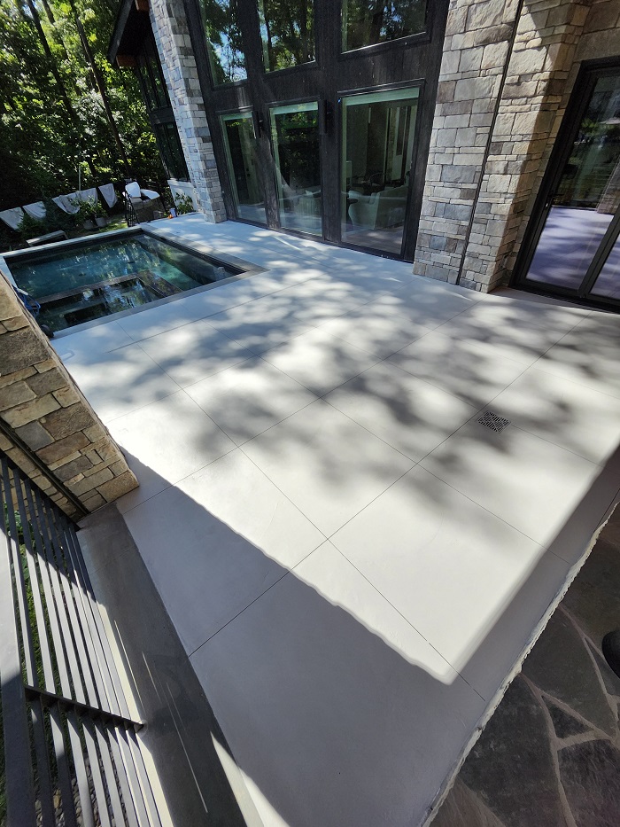 Modern patio with large white tiles next to a small pool, surrounded by stone walls and large windows.