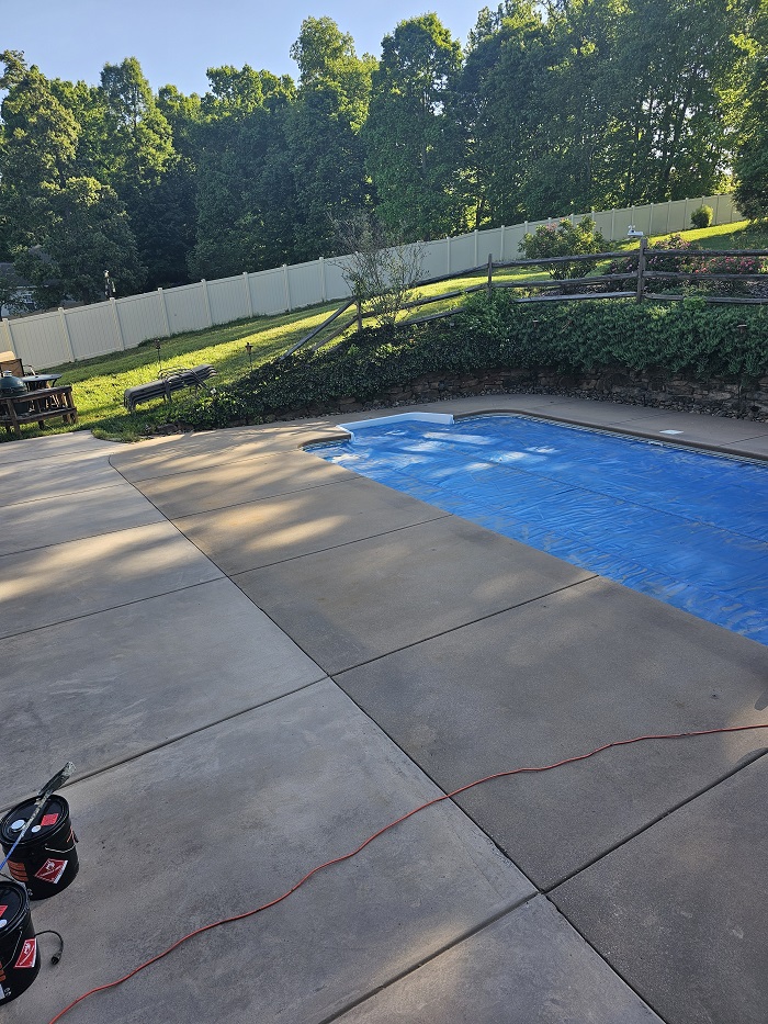 A backyard pool with a blue cover, surrounded by concrete, trees, and a white fence in the background.