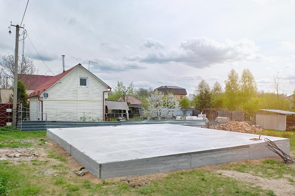 Concrete foundation slab on a grassy plot near a house, with trees and cloudy sky in the background.