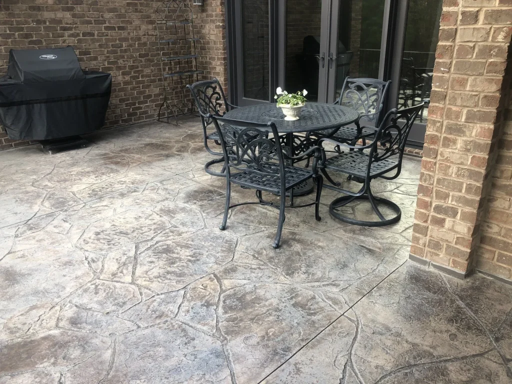 Black metal patio table with four chairs on a stone-patterned patio near a grill and brick wall.