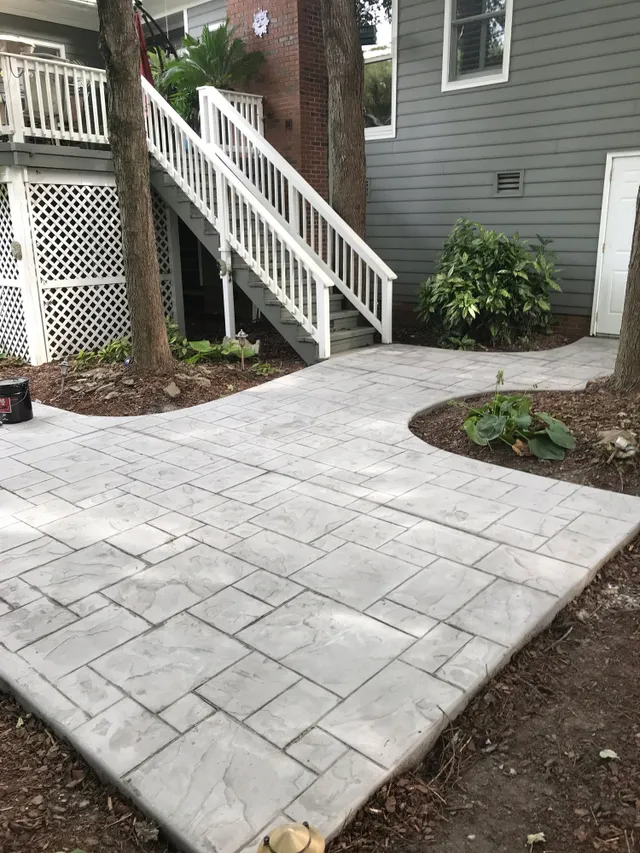 A gray stone patio pathway leads to stairs beside a house, bordered by mulch and plants.