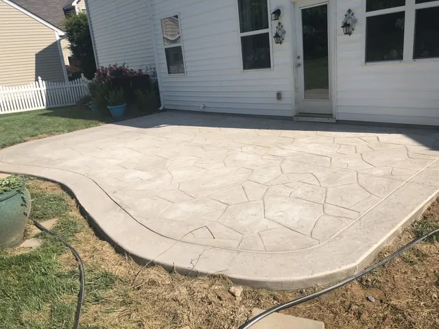 A concrete patio with a decorative pattern is next to a white house and lawn on a sunny day.