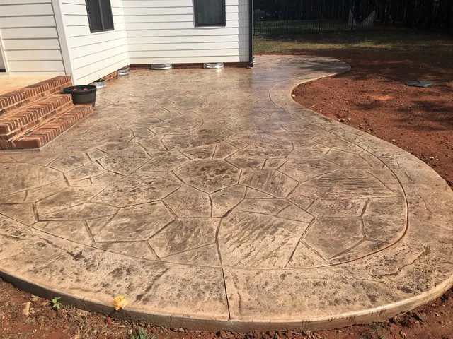 Patio-Waterproofing-By-Youngblood-Waterproofing-Concrete-Services Curved stamped concrete patio with a stone pattern next to a house with brick steps and dirt yard.