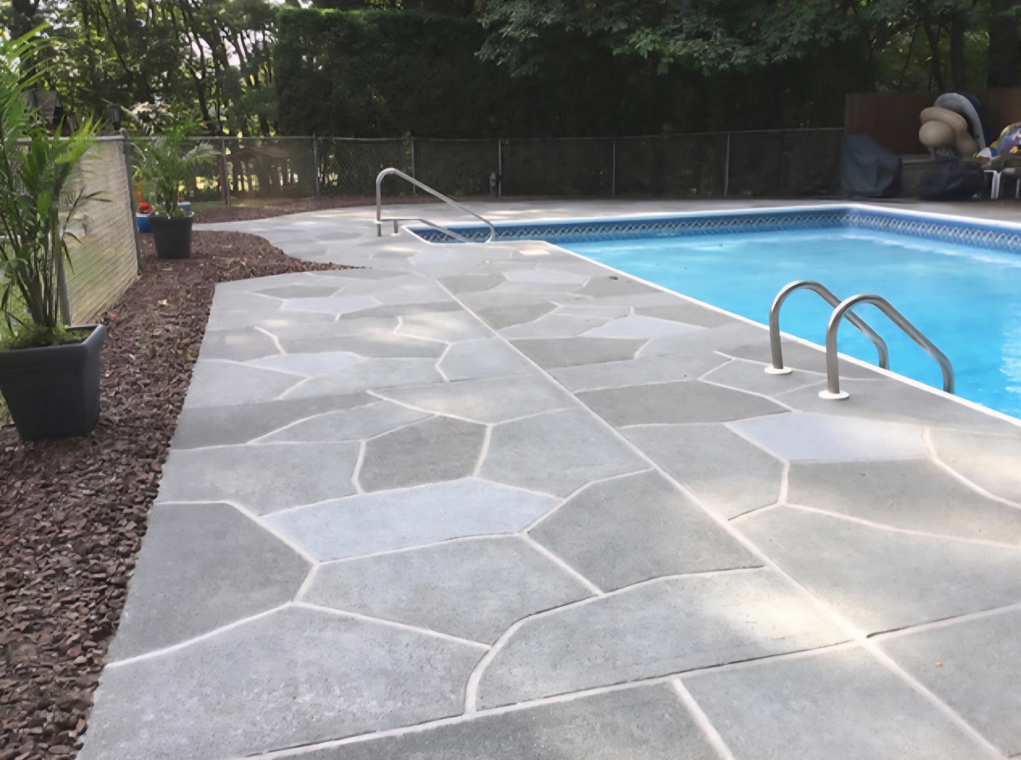 Swimming pool with a geometric patterned concrete deck, surrounded by trees and potted plants.