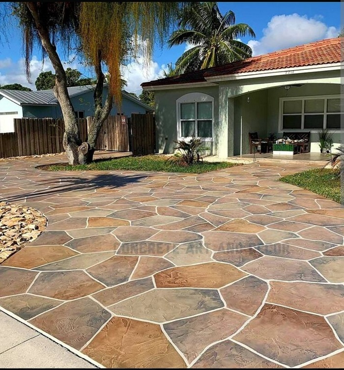 A house with a wide, multi-colored stone-patterned driveway and green lawn, with trees and blue sky.