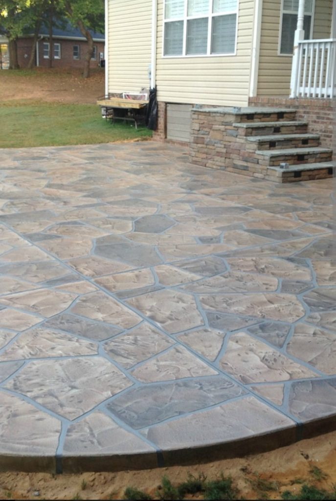 Stamped concrete patio with a stone pattern next to a house with stone steps and a small porch.