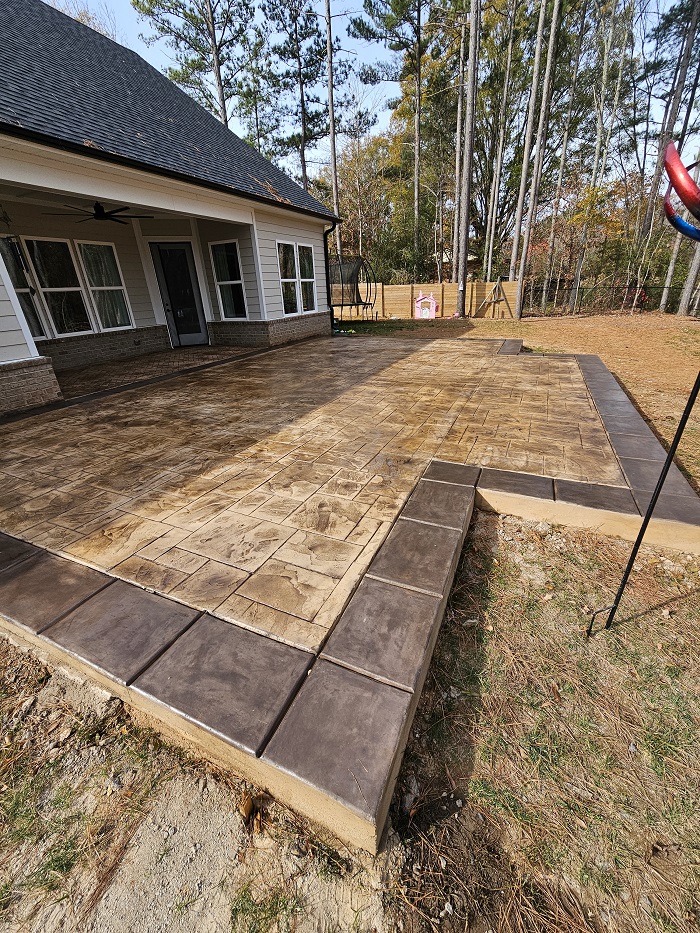 Stamped concrete patio with a brown border beside a house, surrounded by trees and a grassy yard.