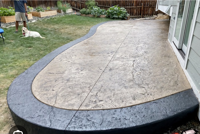 Curved stamped concrete patio with a dark border, next to grass; a dog and person are in the background.