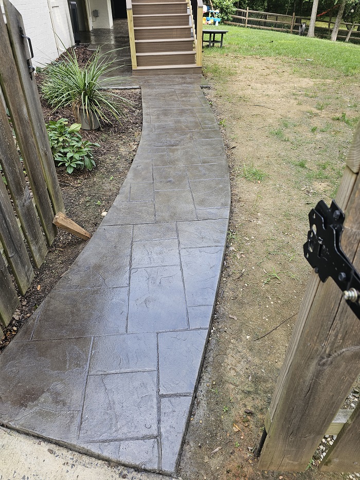 Curved stamped concrete walkway leads to wooden stairs beside a fenced yard and green plants.