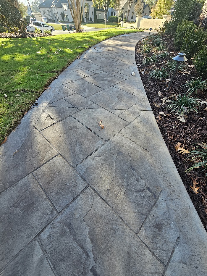Curved concrete walkway bordered by grass and mulch, with houses and trees in the background.