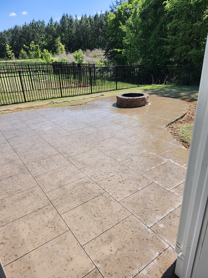 Wet stone patio with a round fire pit, black metal fence, and trees in the background under a blue sky.