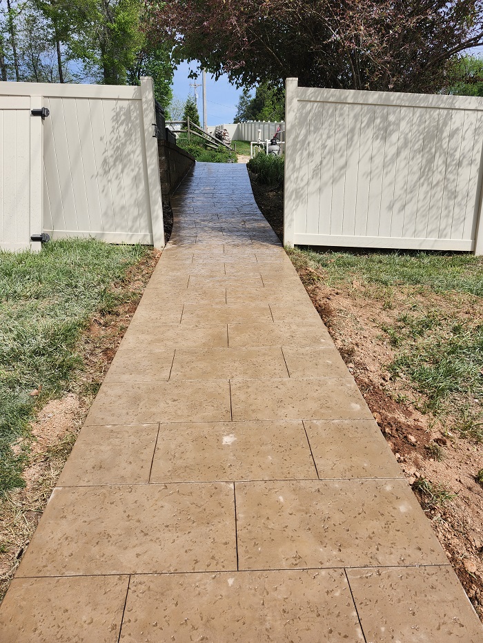 A tan, tiled walkway leads through an open white gate toward a sunny, tree-lined yard.