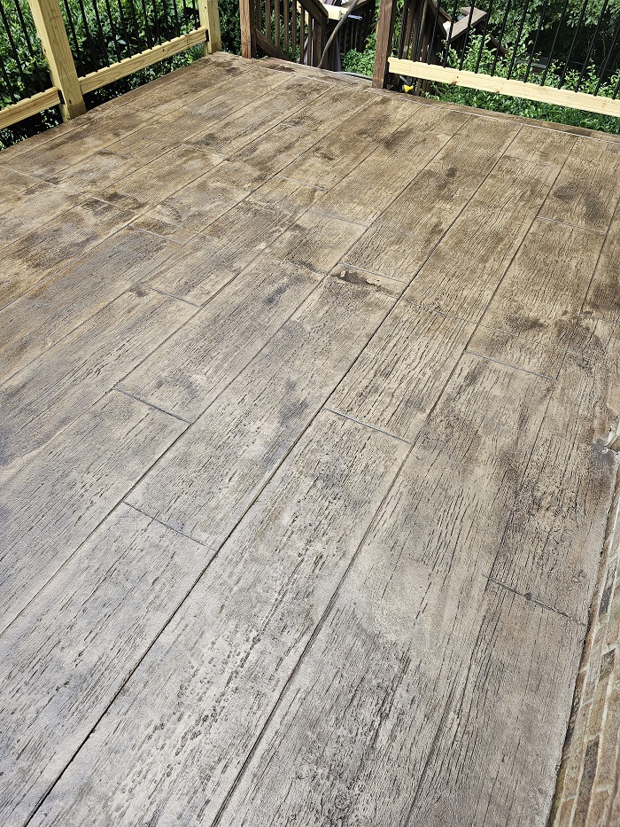 Wood-textured concrete patio with a plank pattern, surrounded by a wooden railing and greenery.