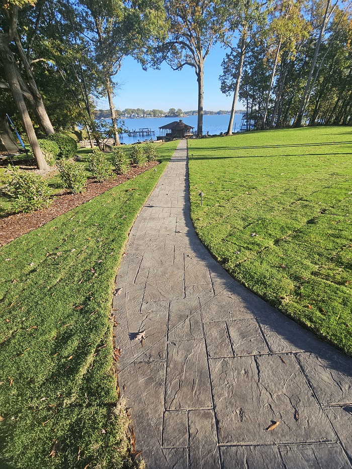 A stone path leads through a green lawn toward a dock and lake, surrounded by trees on a sunny day.