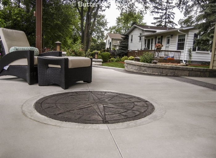 A patio with a compass design stamped into the concrete, outdoor chairs, and a house with a garden.