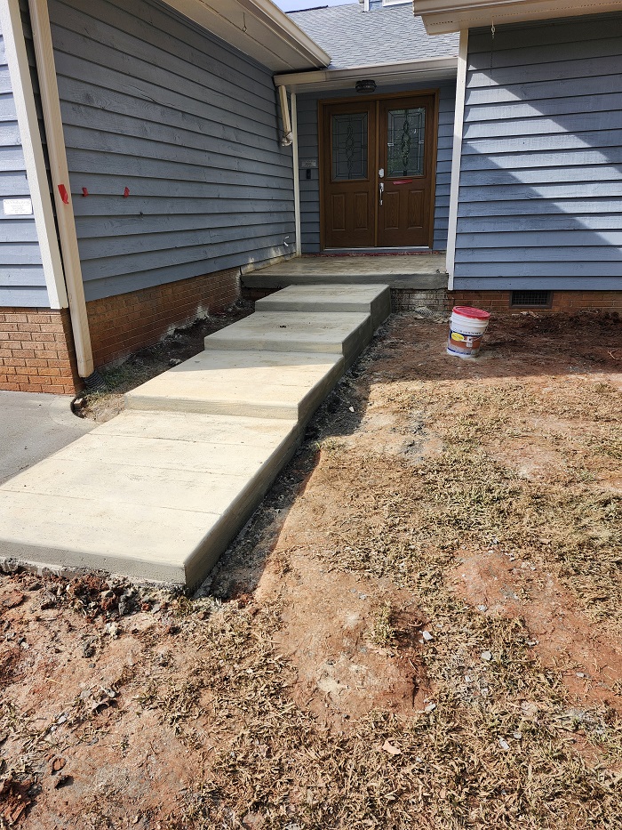Concrete sidewalk leads to a home's front steps; yard is bare dirt with a red and white bucket on the right.