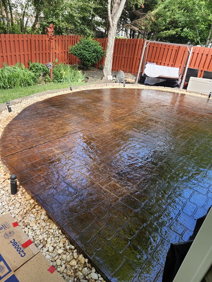 Stamped concrete patio with wet surface, bordered by rocks, in a fenced backyard with trees and garden.