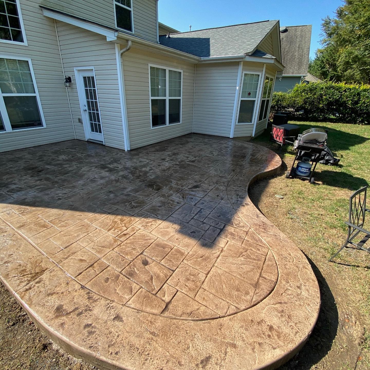 Stamped concrete patio with curved edges next to a house, with a grill and chair on the lawn.