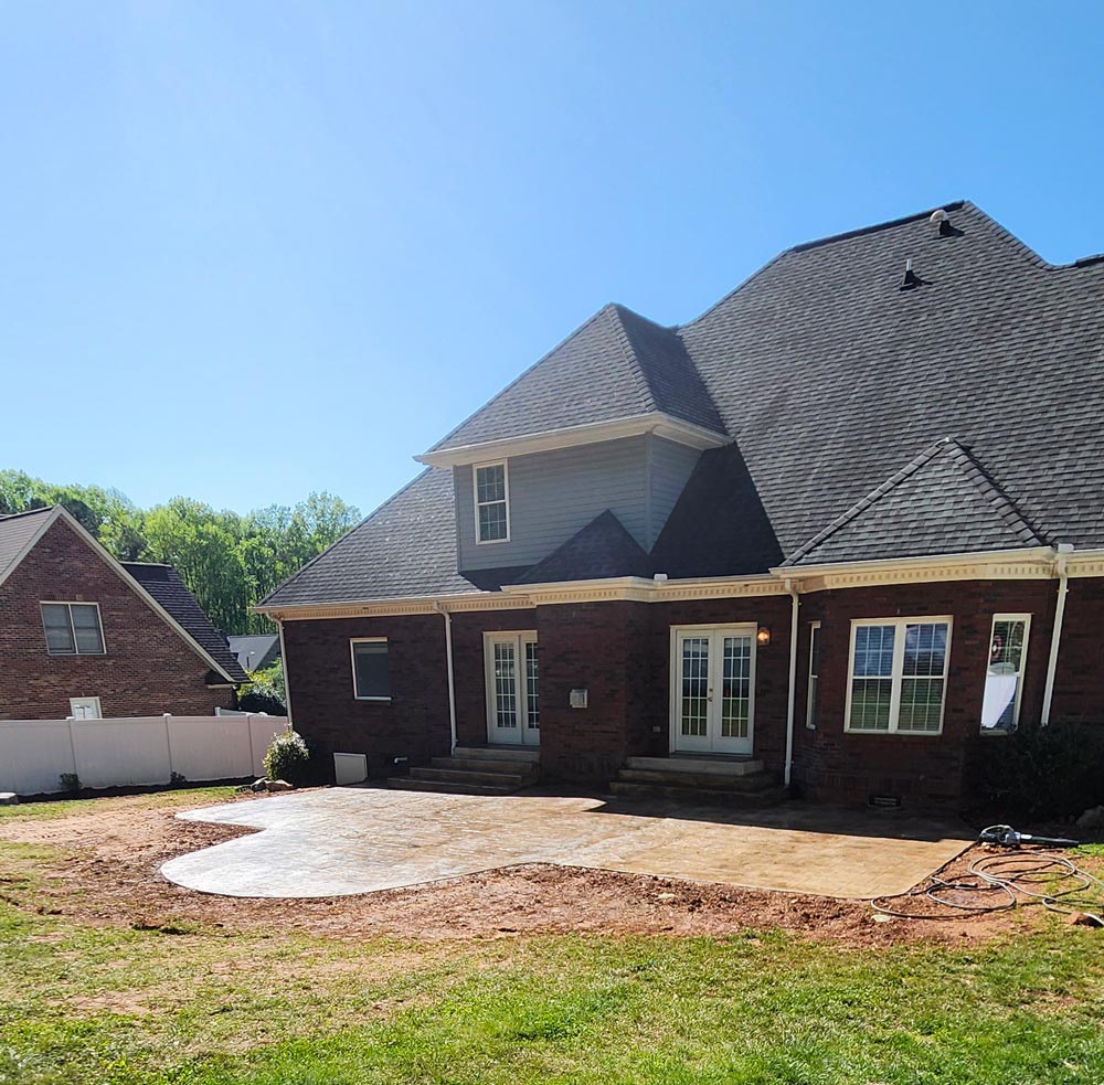 A newly poured concrete patio extends from the back of a brick house under a clear blue sky.