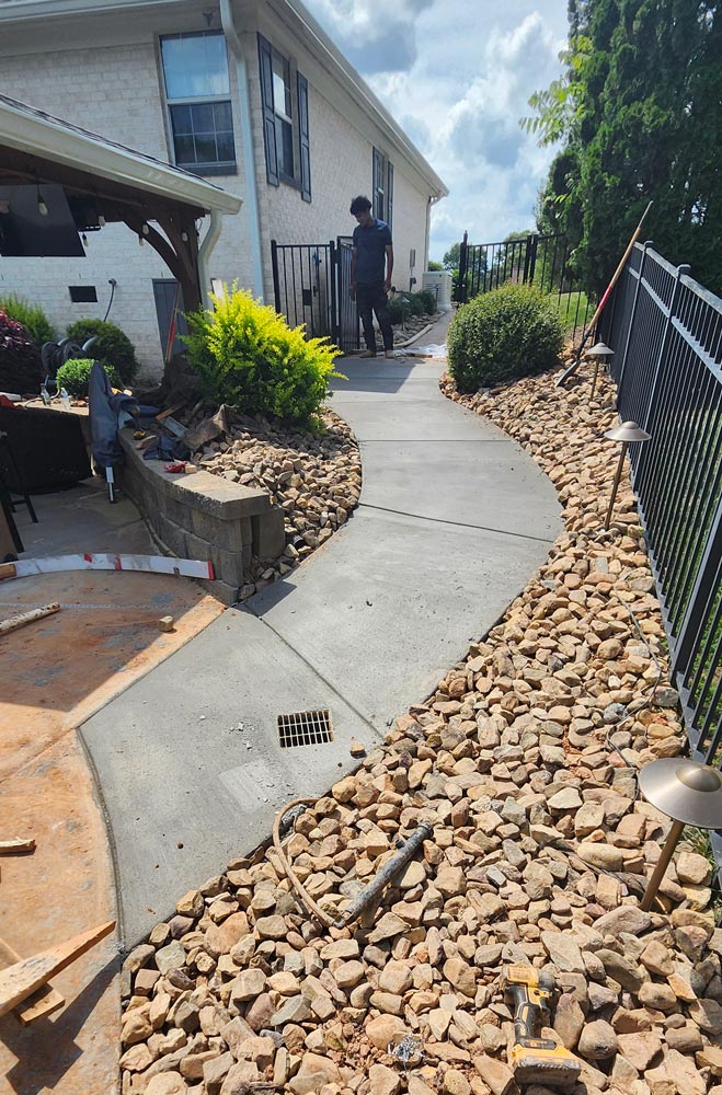 Curved concrete walkway bordered by rocks, with a person standing near a black fence by a house.