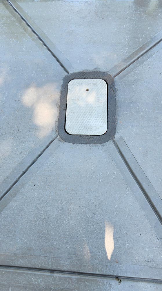 A small metal utility cover is set in a gray concrete surface with intersecting lines.