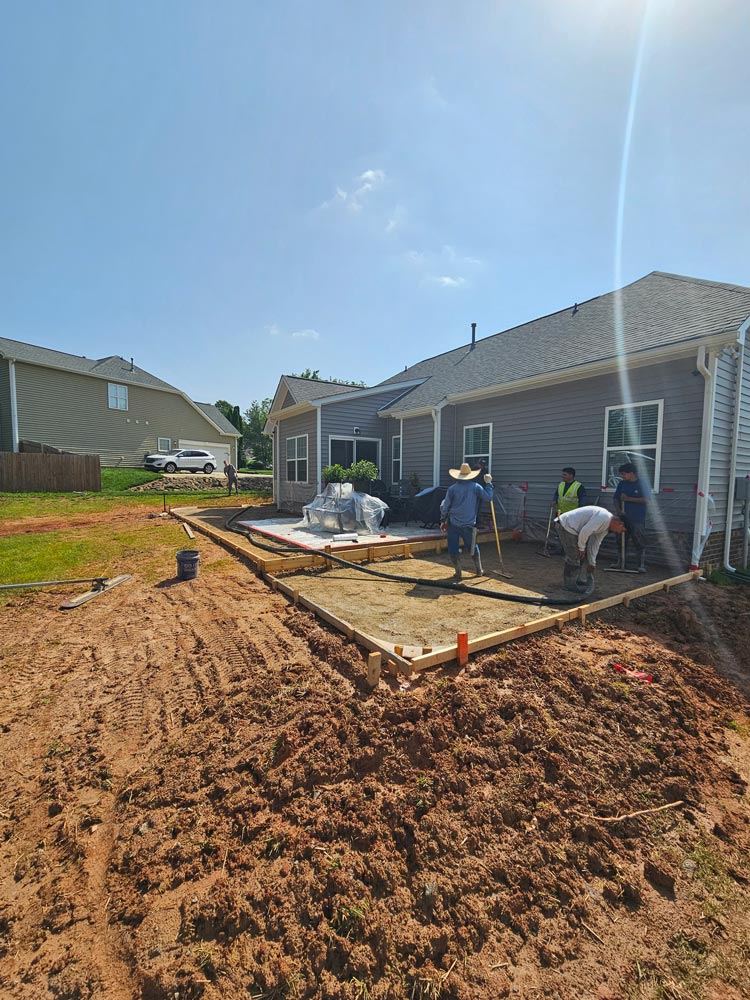 Four workers prepare a backyard for a concrete patio beside a gray house under a clear blue sky.