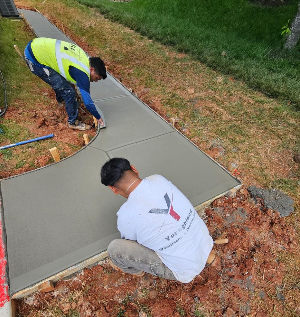 Two workers smooth freshly poured concrete on a curved sidewalk near grass and dirt.