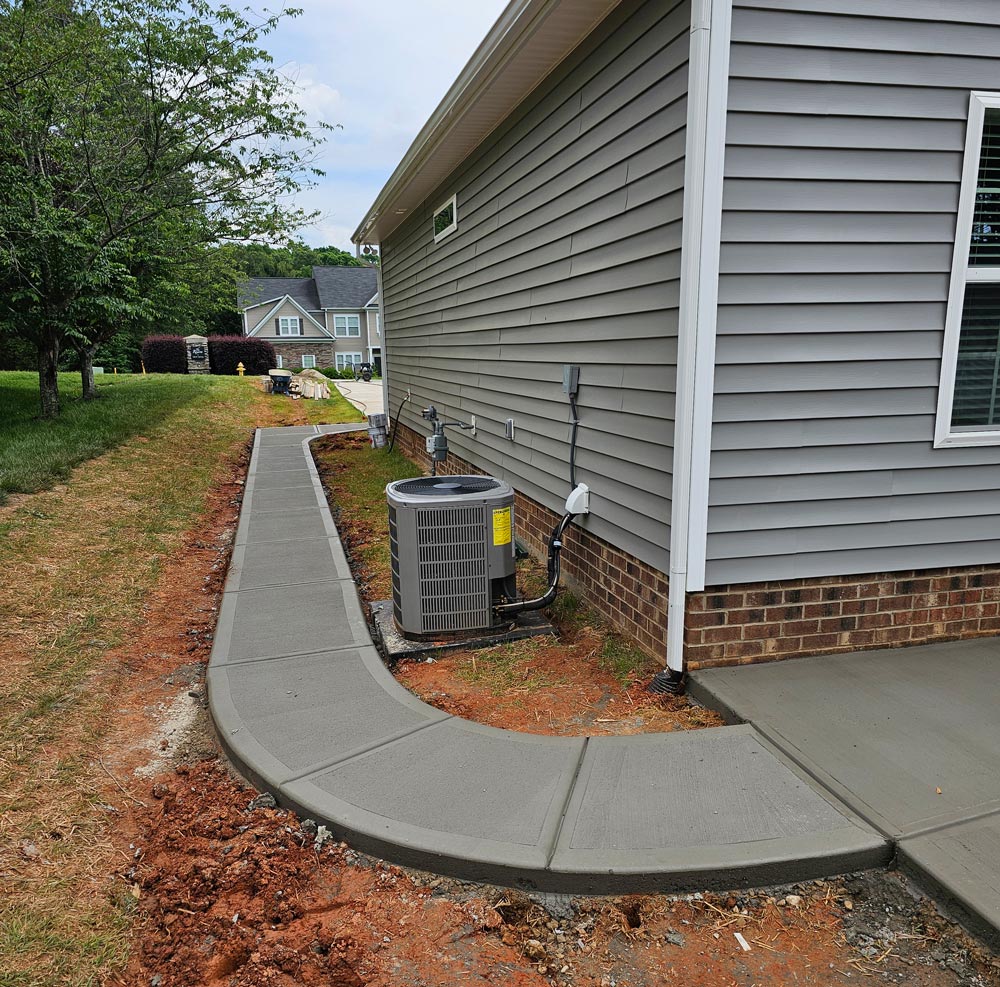 Freshly poured concrete sidewalk curves around the side of a gray house with a brick foundation.