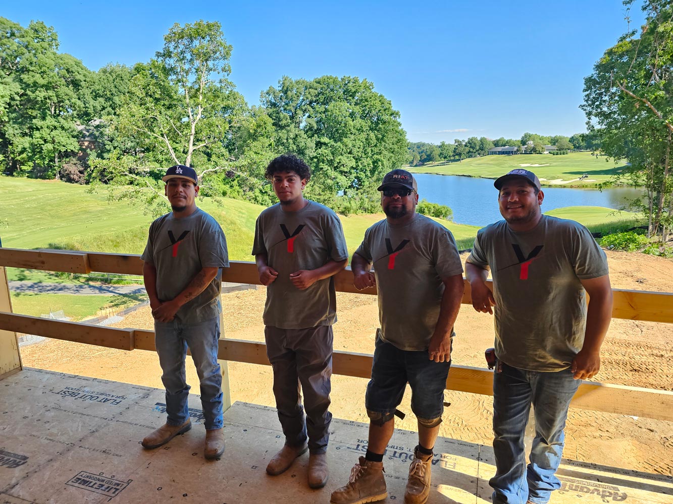 Four men in matching t-shirts stand on a balcony overlooking a green landscape with trees and a pond.
