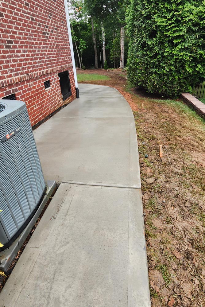 Freshly poured concrete walkway curves alongside a brick house, bordered by grass and trees.