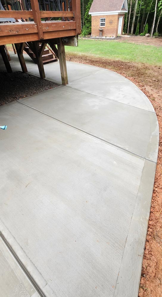 Freshly poured curved concrete walkway next to a wooden deck, with grass and a small shed in the background.