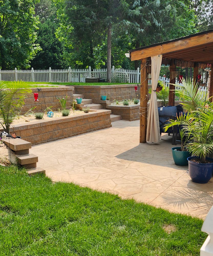 A sunny backyard patio with potted plants, stone steps, and a covered seating area next to a grassy lawn.