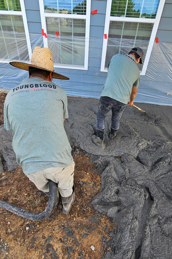 Two workers in gray shirts pour and level concrete outside a house with plastic-covered windows.