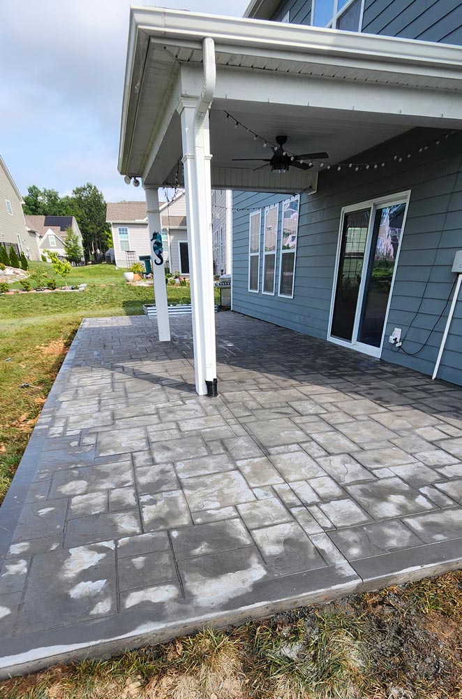 Newly installed gray stone patio with white grout, beneath a covered porch attached to a blue house.