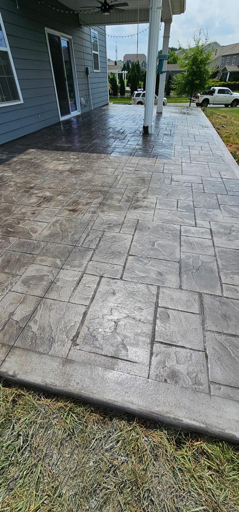 Stamped concrete patio with a square tile pattern, next to a gray house and grassy yard.